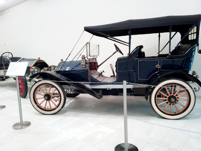 [Side view of the car with a black roof and open on the front and both sides. There are no front doors, but there are doors for the rear seat. The wooden spoke wheels have white-walled tires. The column for the steering wheel is very long to reach the high seat of the driver.]